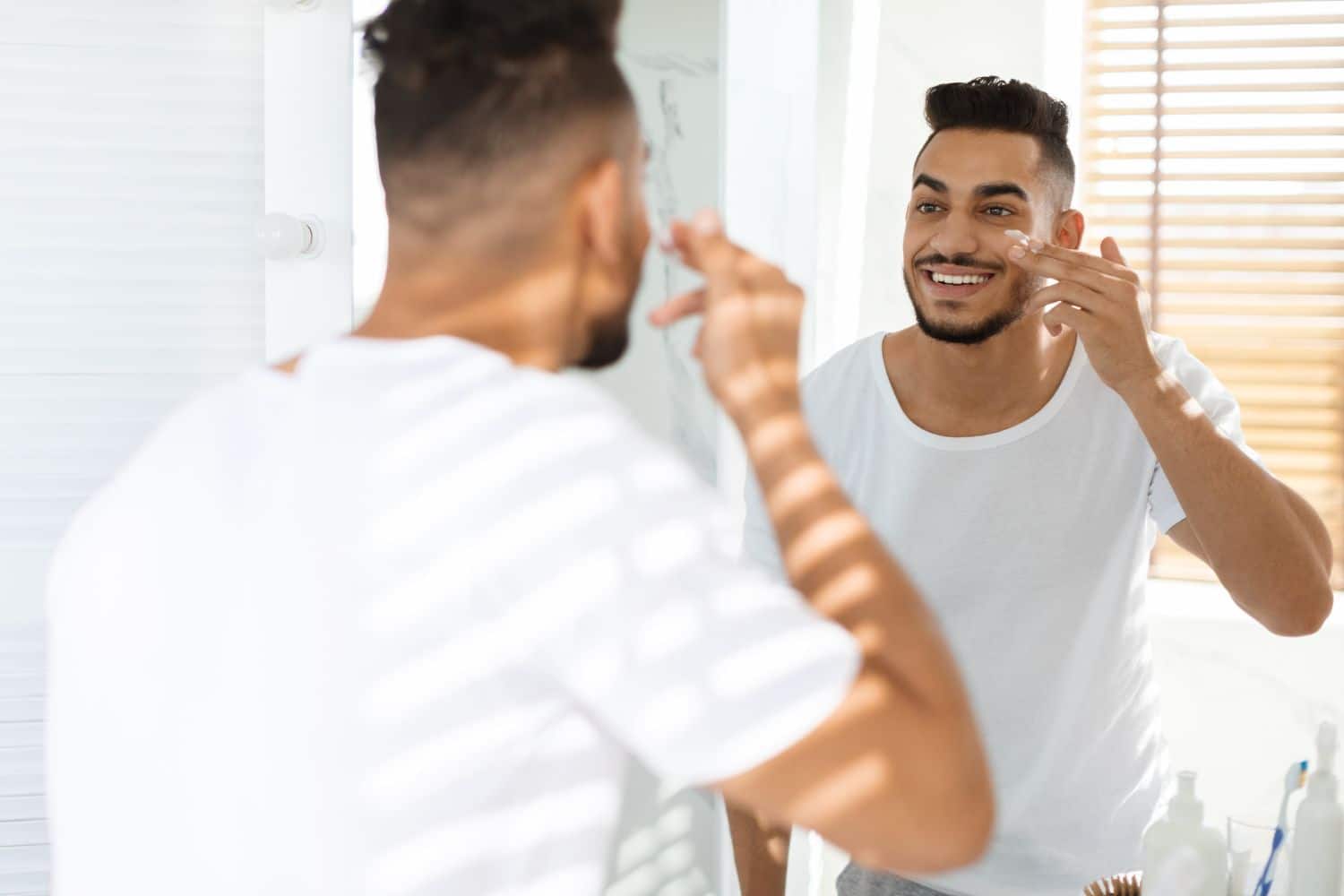 man rubbing face cream just below eyes in front of mirror